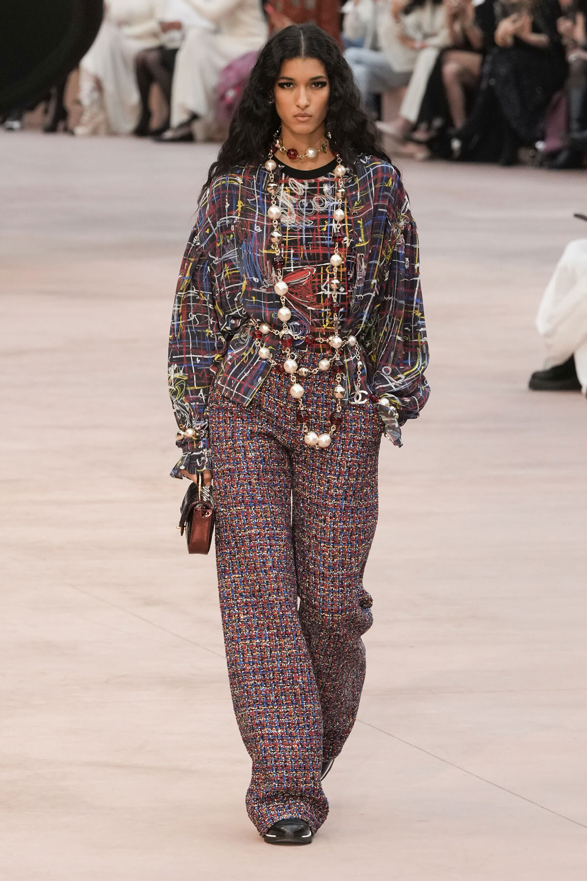 Model walks runway in a loose, multicolored plaid top with matching printed wide-leg pants and layered pearl necklaces, carrying a small burgundy handbag.