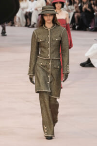 Model walks the runway in a textured olive tweed suit with braided trim and matching hat.