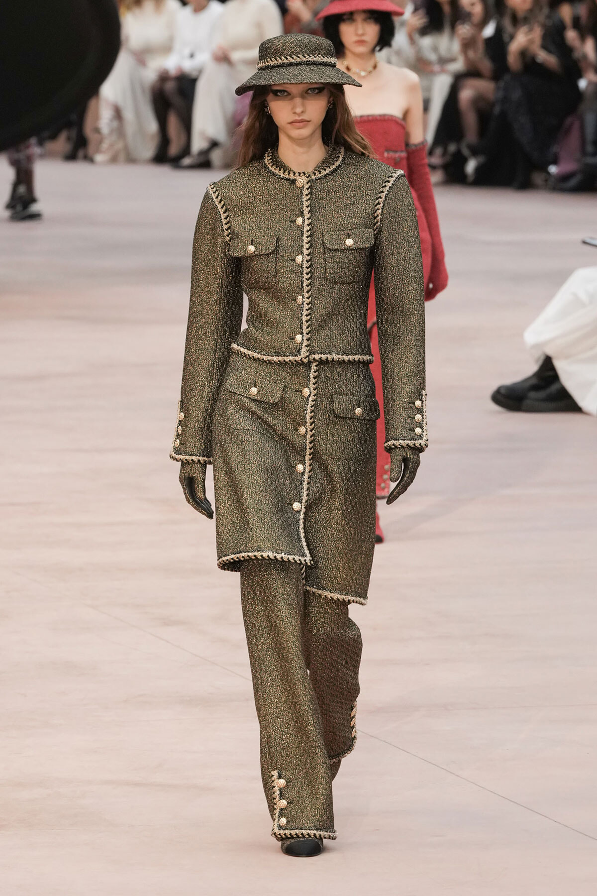 Model walks the runway in a textured olive tweed suit with braided trim and matching hat.