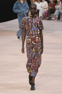 Model walking a runway in a multicolor graffiti-print dress with a long pearl necklace and a small metallic crossbody bag.