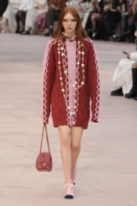 Model walking runway in pink and burgundy cable-knit dress with layered pearl necklaces and a pink tweed handbag.