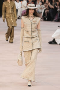 Runway model in a cream tweed ensemble (sleeveless jacket and wide-leg pants) with a matching hat, gloves, and handbag.