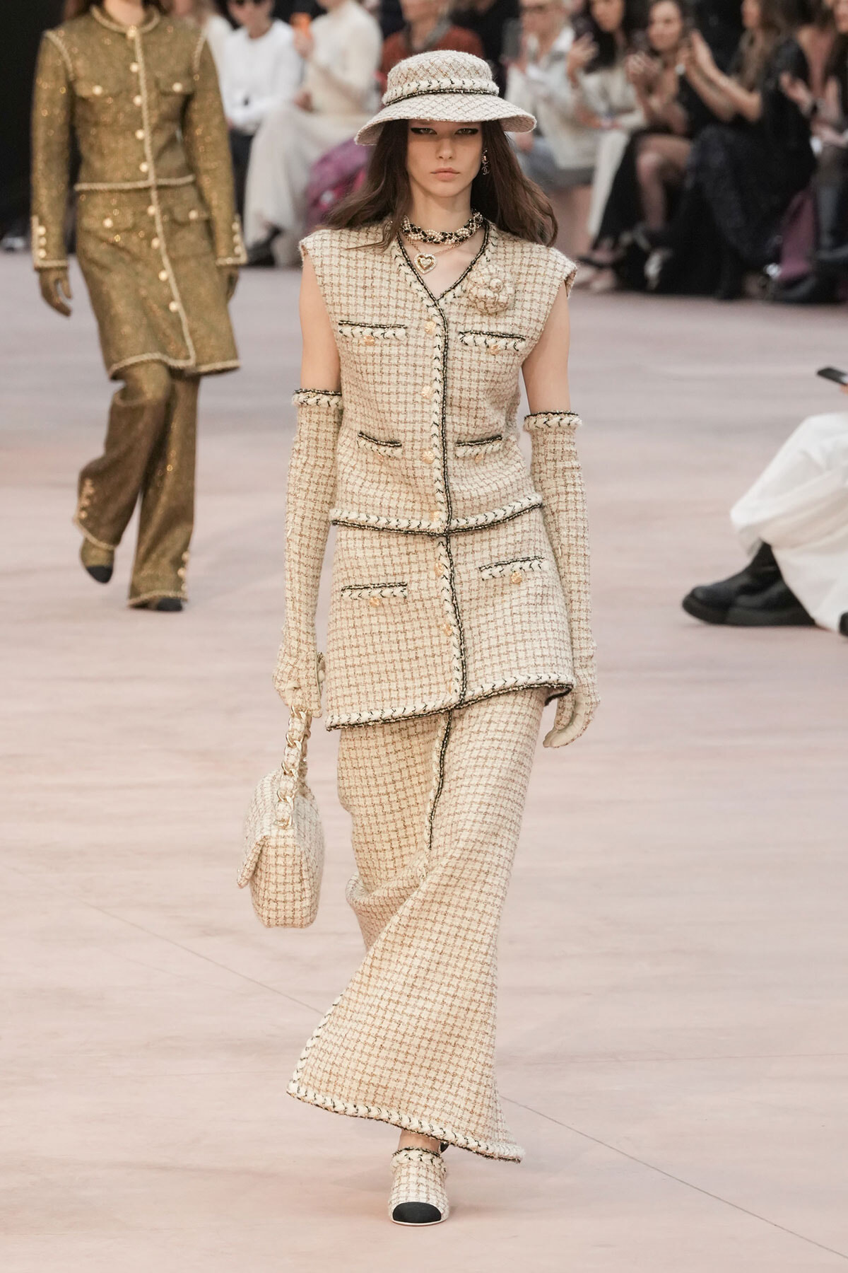 Runway model in a cream tweed ensemble (sleeveless jacket and wide-leg pants) with a matching hat, gloves, and handbag.