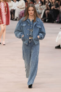 Model walks the runway in a blue corduroy matching jacket and wide-leg pants, accessorized with layered silver jewelry and hands in pockets.
