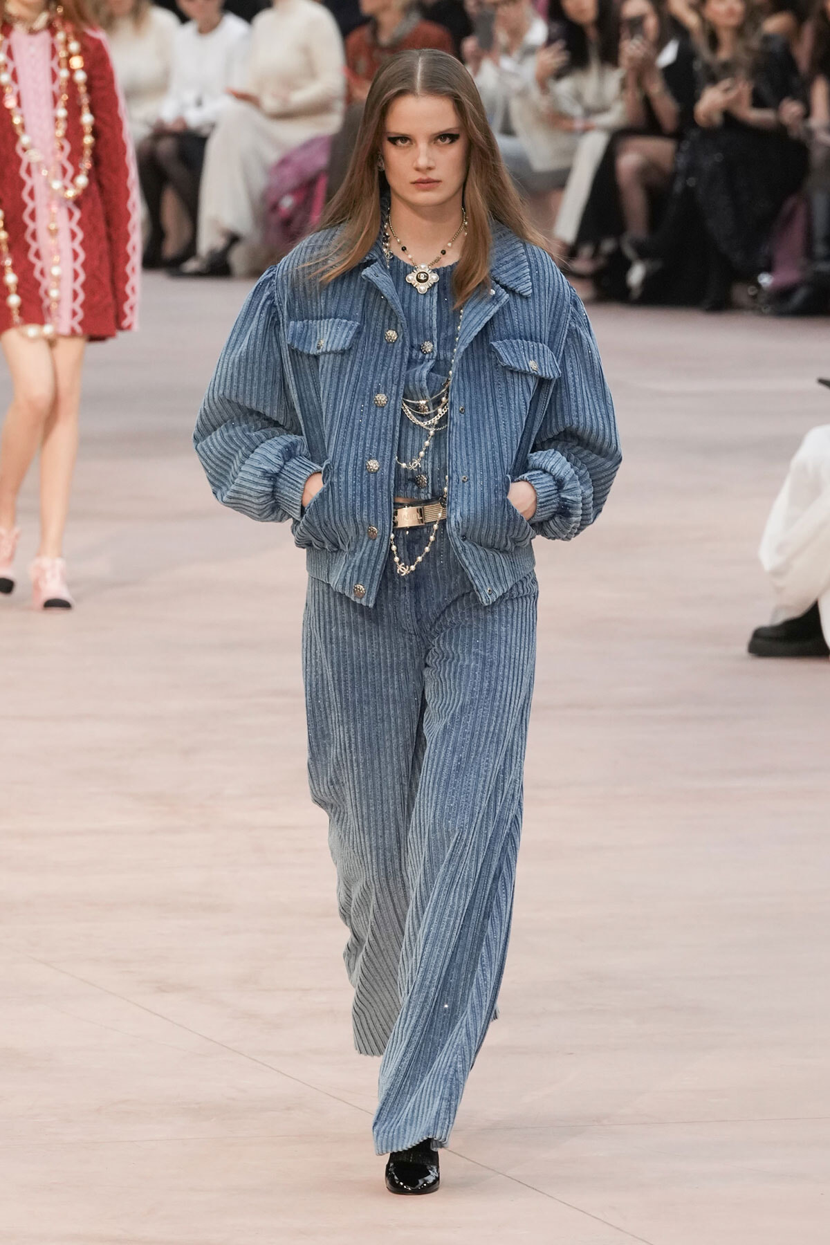Model walks the runway in a blue corduroy matching jacket and wide-leg pants, accessorized with layered silver jewelry and hands in pockets.