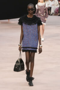 Model walking runway in a blue tweed shift dress with black ruffled collar and cuffs, holding a quilted black handbag and wearing oversized sunglasses.