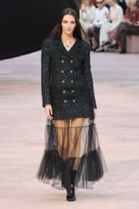 Model walking the runway in a black tweed double-breasted jacket with matching mini skirt and a sheer black tulle overlay.