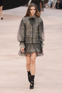 Model walking a runway in a tweed two-piece set with sheer black puff sleeves and a tulle-trimmed mini skirt, black bow at the neck.