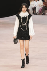 Model on a runway wearing a black tweed mini dress over a white puff-sleeve blouse with a ruffled collar, layered pearl necklaces, and black boots, carrying a studded clutch.