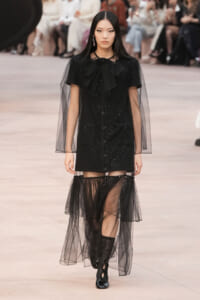 Model walking the runway in a black embellished mini dress with sheer cape sleeves and a layered tulle overlay skirt.