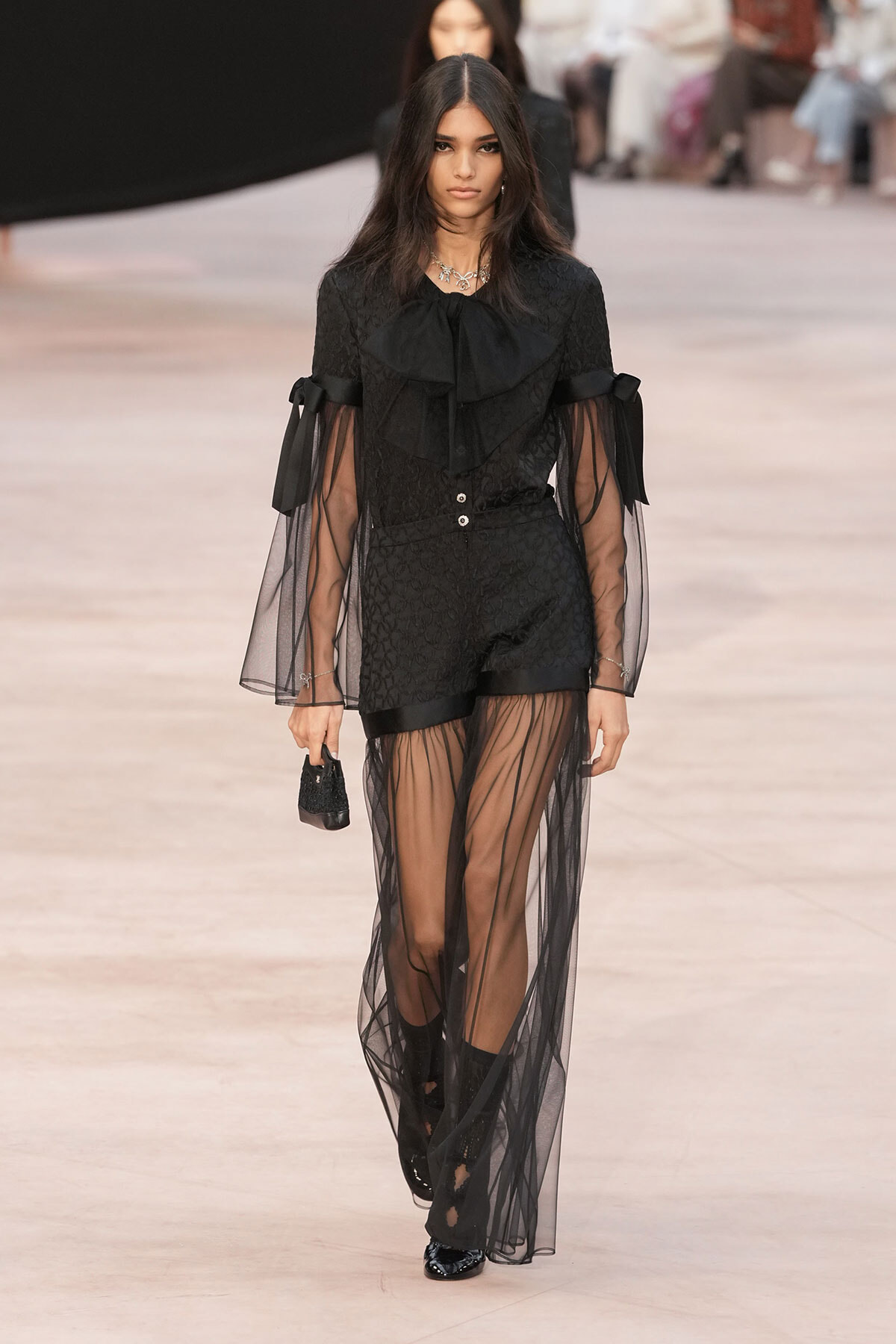 Runway model in a black lace outfit with sheer overlay and large bow at the neck.