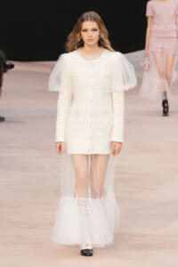 Runway model in a white textured coat dress with a sheer tulle cape draping from the shoulders to the floor.