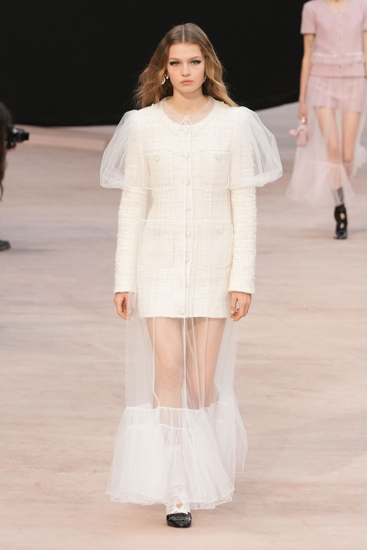 Runway model in a white textured coat dress with a sheer tulle cape draping from the shoulders to the floor.