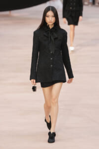 Model walking runway in a black sequined blazer dress with a large bow necktie and matching mini skirt, carrying a tiny handbag.