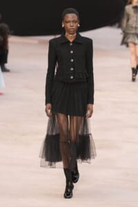 Model on a runway wearing a black textured button-front jacket and a sheer, layered tulle skirt with black shoes.