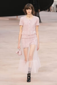Runway model in pastel pink tweed set with sheer tulle overlay, holding a pink handbag and wearing a choker necklace.