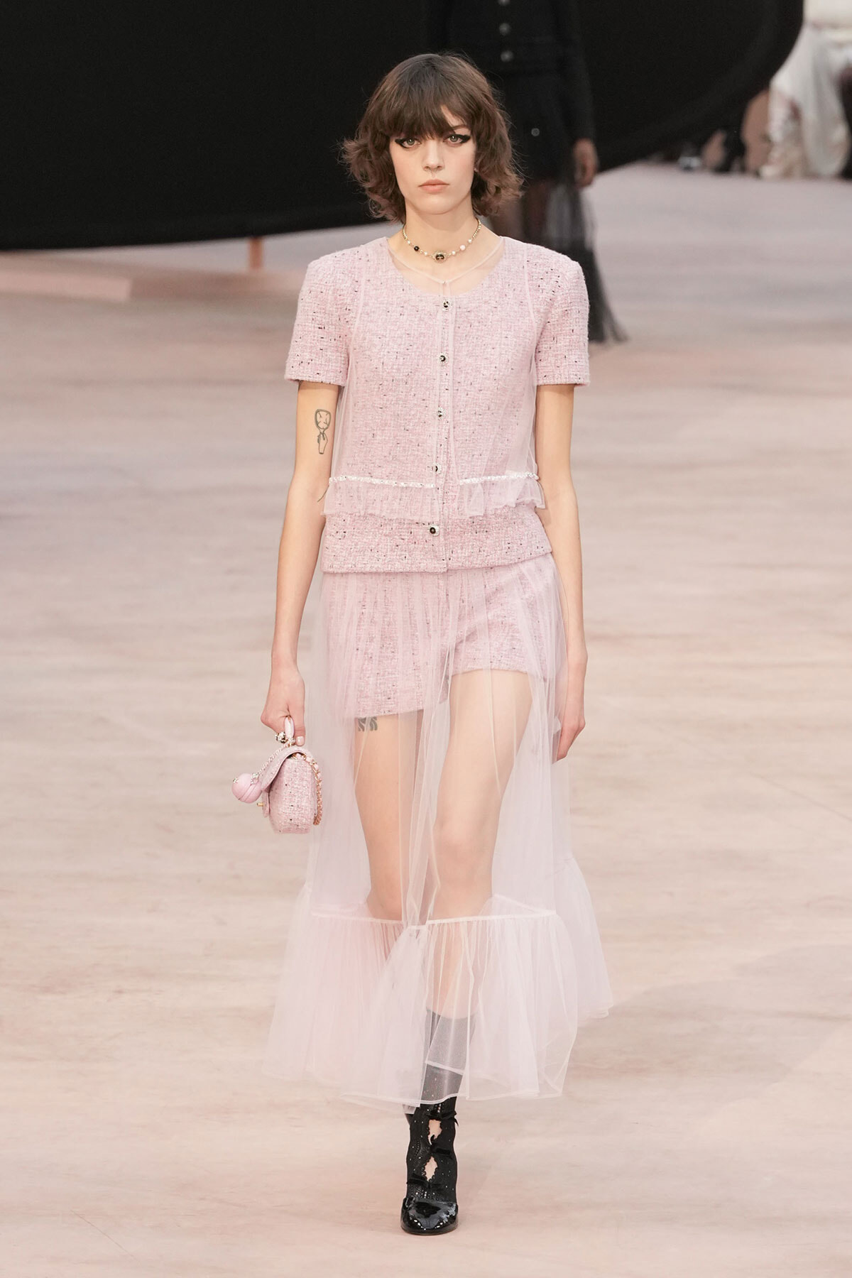 Runway model in pastel pink tweed set with sheer tulle overlay, holding a pink handbag and wearing a choker necklace.