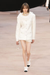 Model walking a runway in a white tweed blazer and shorts with a ruffled collar and chunky necklace, carrying a small spherical bag on a chain.