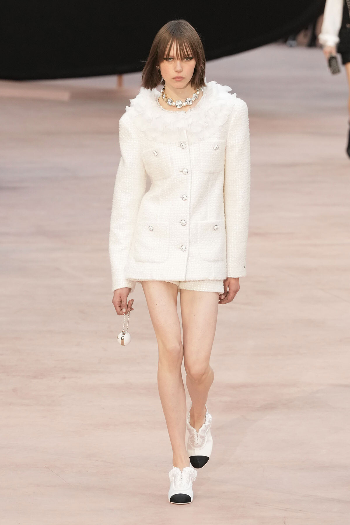 Model walking a runway in a white tweed blazer and shorts with a ruffled collar and chunky necklace, carrying a small spherical bag on a chain.