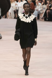 Model walking the runway in a black sweater dress with a large white floral collar, holding a textured coat in one arm.