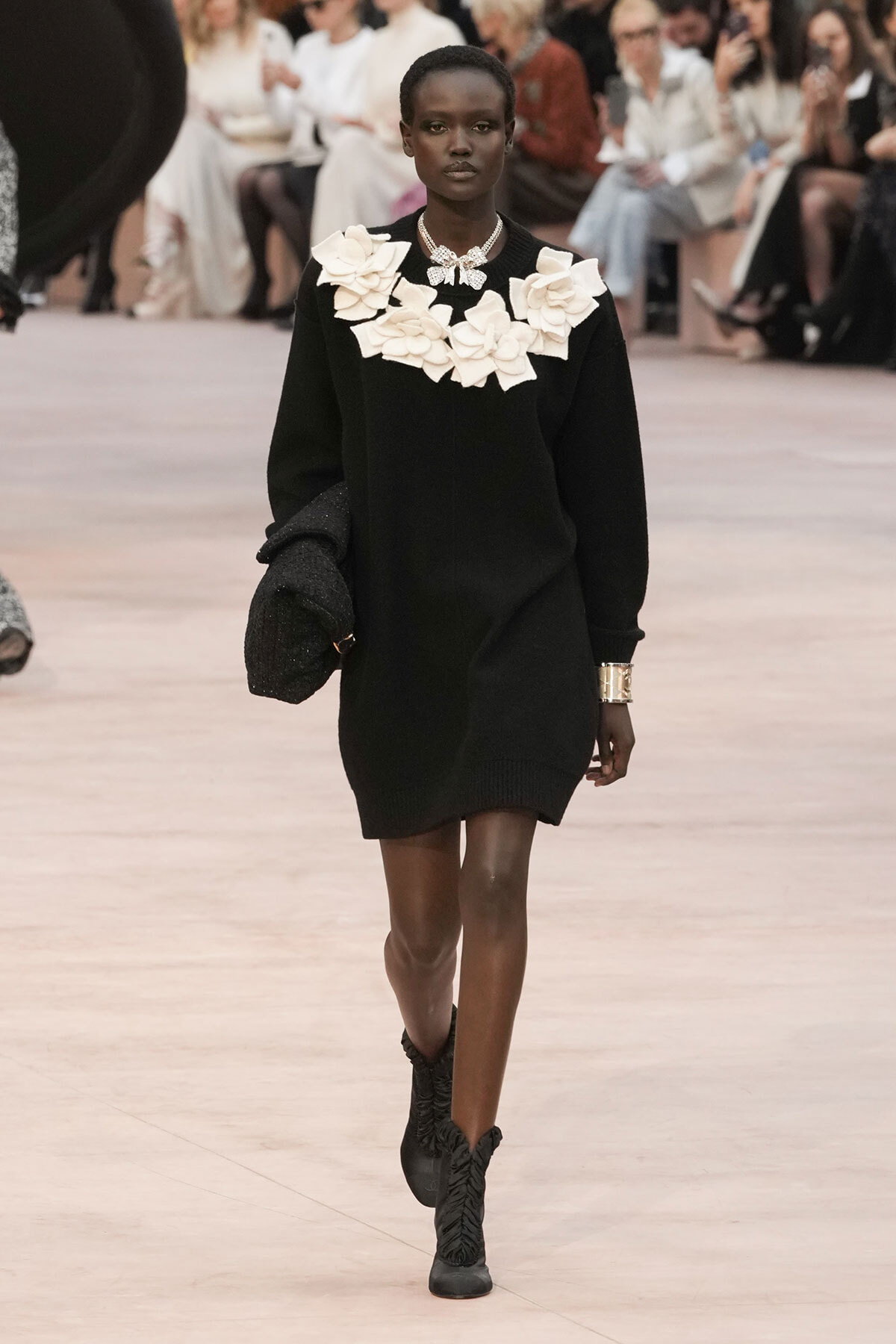 Model walking the runway in a black sweater dress with a large white floral collar, holding a textured coat in one arm.
