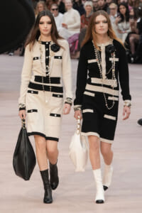 Two models walk a fashion runway in coordinated beige and black knit dresses with pockets, layered pearl necklaces, bracelets, and oversized handbags; audience in the background.