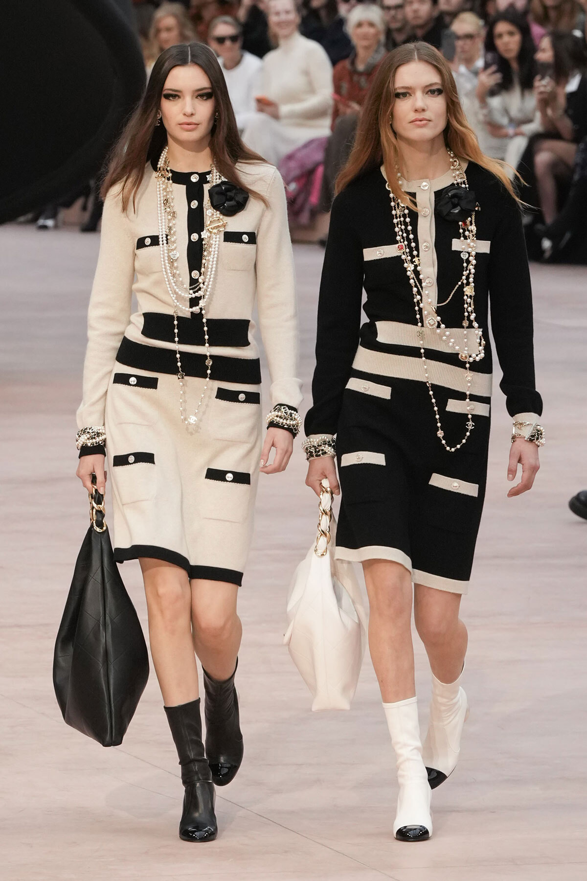 Two models walk a fashion runway in coordinated beige and black knit dresses with pockets, layered pearl necklaces, bracelets, and oversized handbags; audience in the background.
