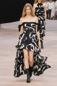 Runway model in a black-and-white abstract print off-the-shoulder dress with bows at the arms and a flowing cape-like train, paired with black boots.