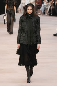 Model walking a runway in a textured black tweed jacket with black ribbon tie and a layered black skirt, paired with knee-high boots.