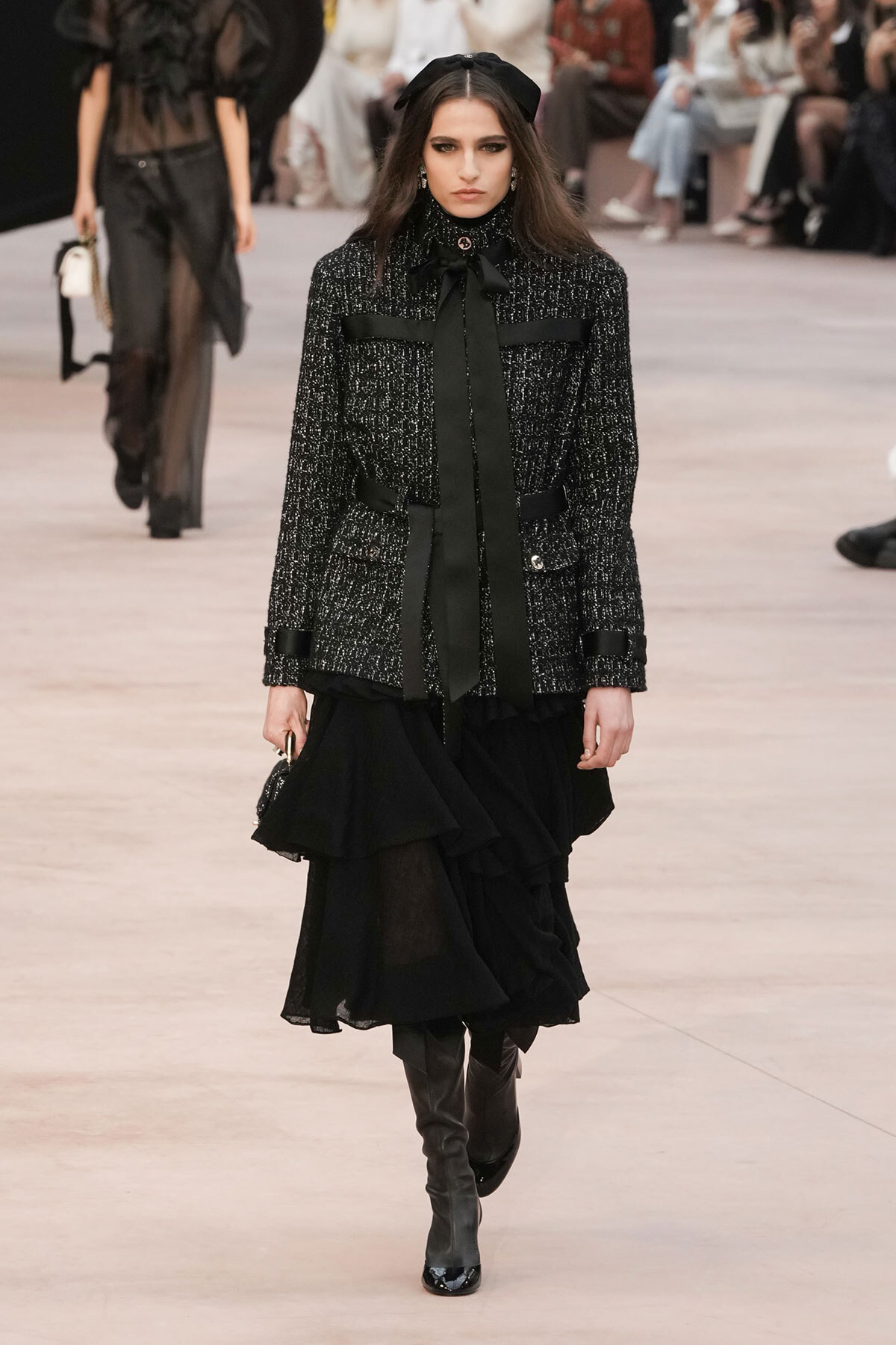 Model walking a runway in a textured black tweed jacket with black ribbon tie and a layered black skirt, paired with knee-high boots.