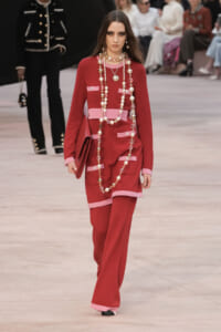 Model walking the runway in a red dress with pink trim and long pearl necklaces, carrying a red clutch in a fashion show setting.