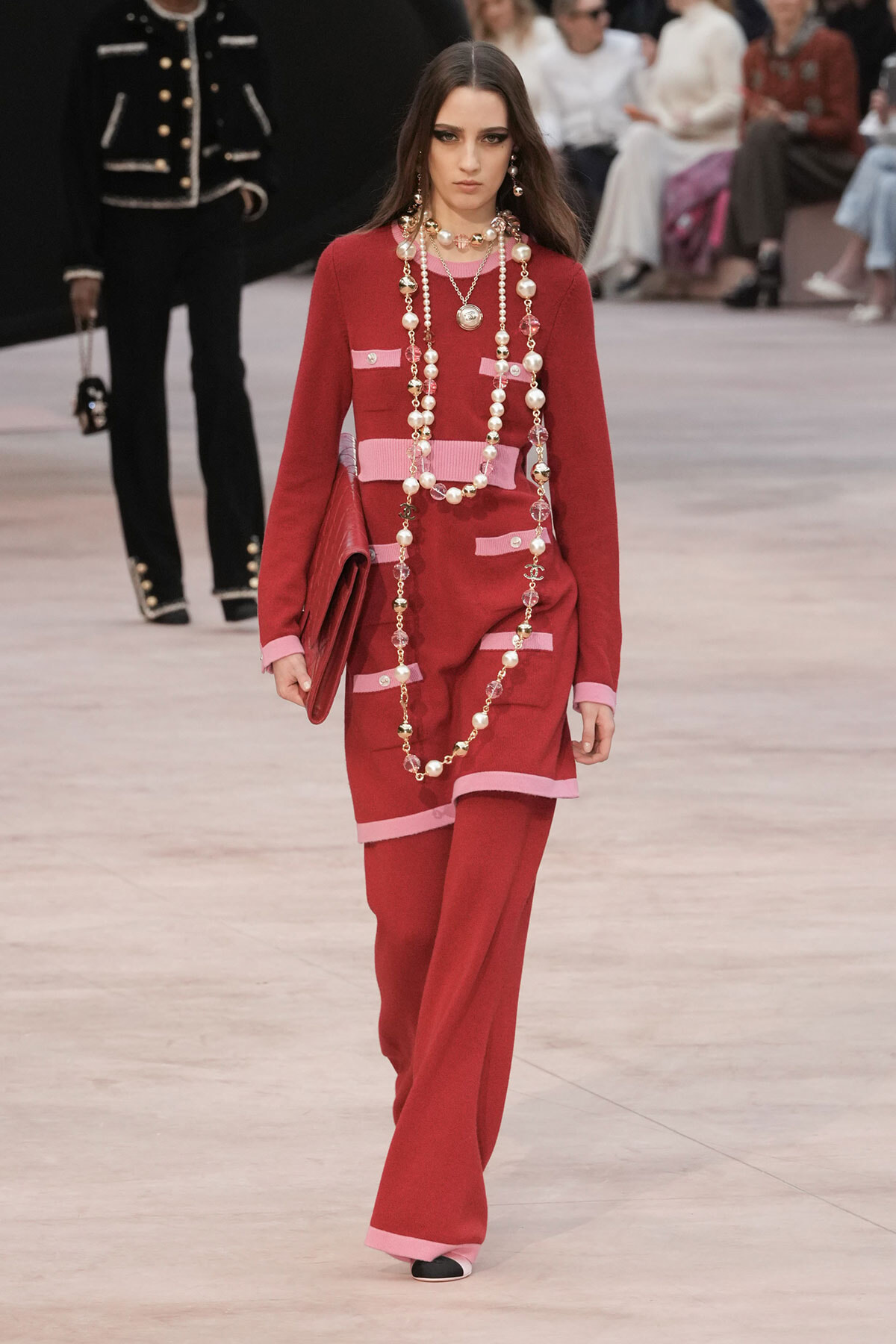 Model walking the runway in a red dress with pink trim and long pearl necklaces, carrying a red clutch in a fashion show setting.