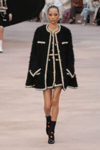 Model walks the runway in a knee-length black bouclé coat with cream trim and gold buttons, accessorized with jewelry.