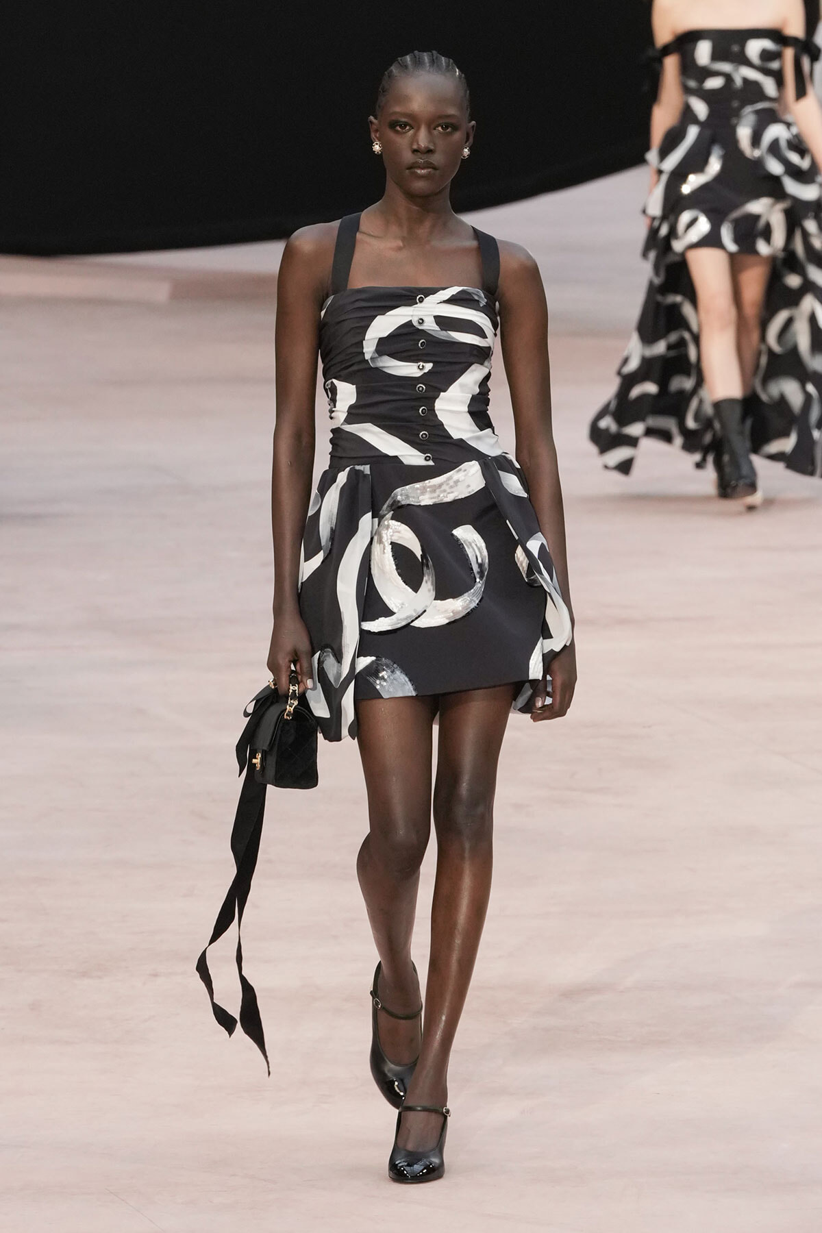 Model walks the runway in a black-and-white abstract-print mini dress with broad straps, carrying a small black handbag.