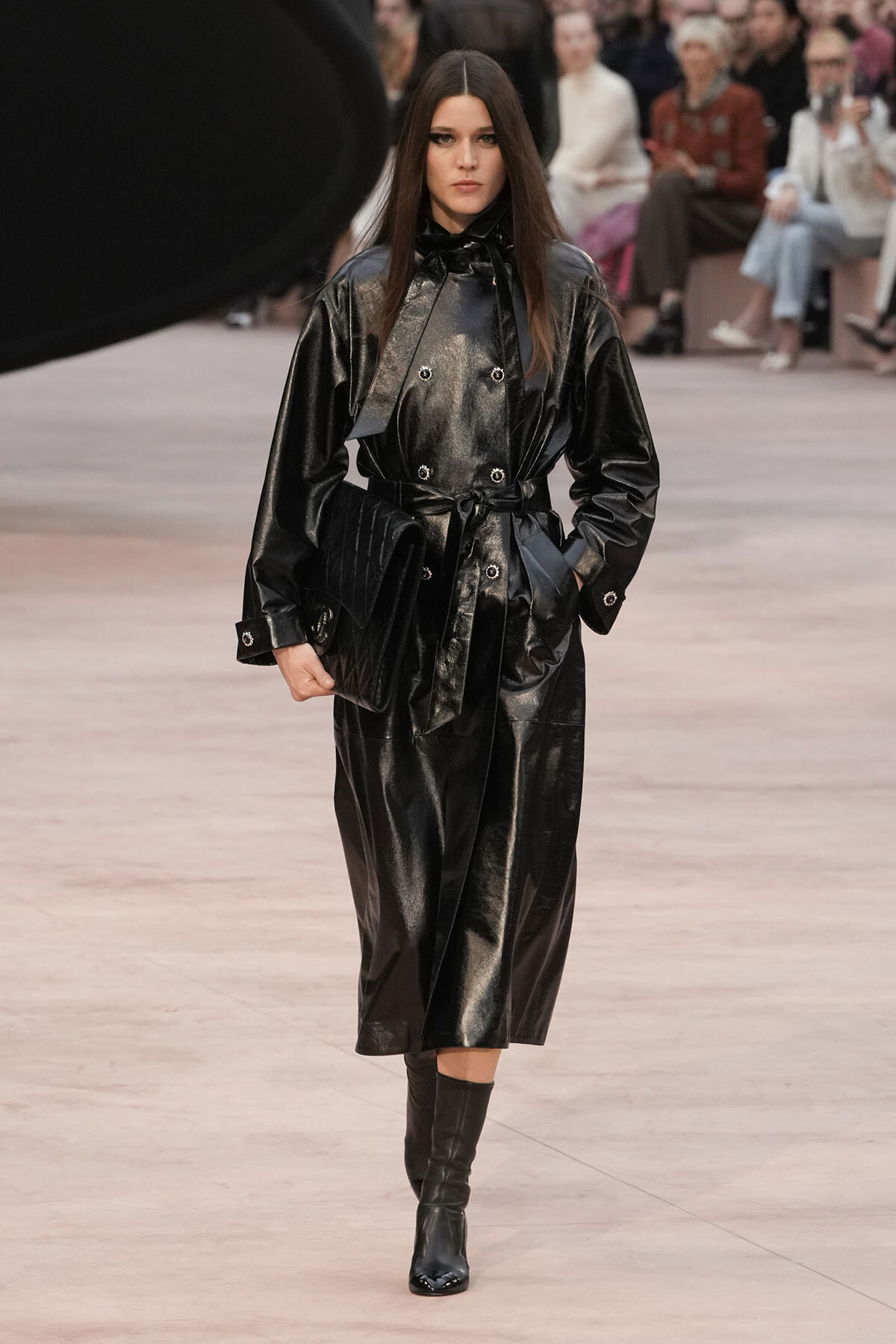 Model walking the runway in a glossy black trench coat with a belt and knee-high boots.