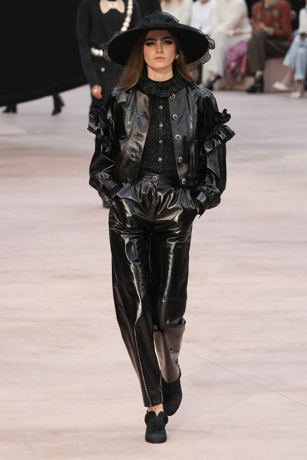 Model walking a fashion show in a black patent-leather suit with puffed sleeves and a wide-brim hat, wearing a sparkly black top and black shoes.