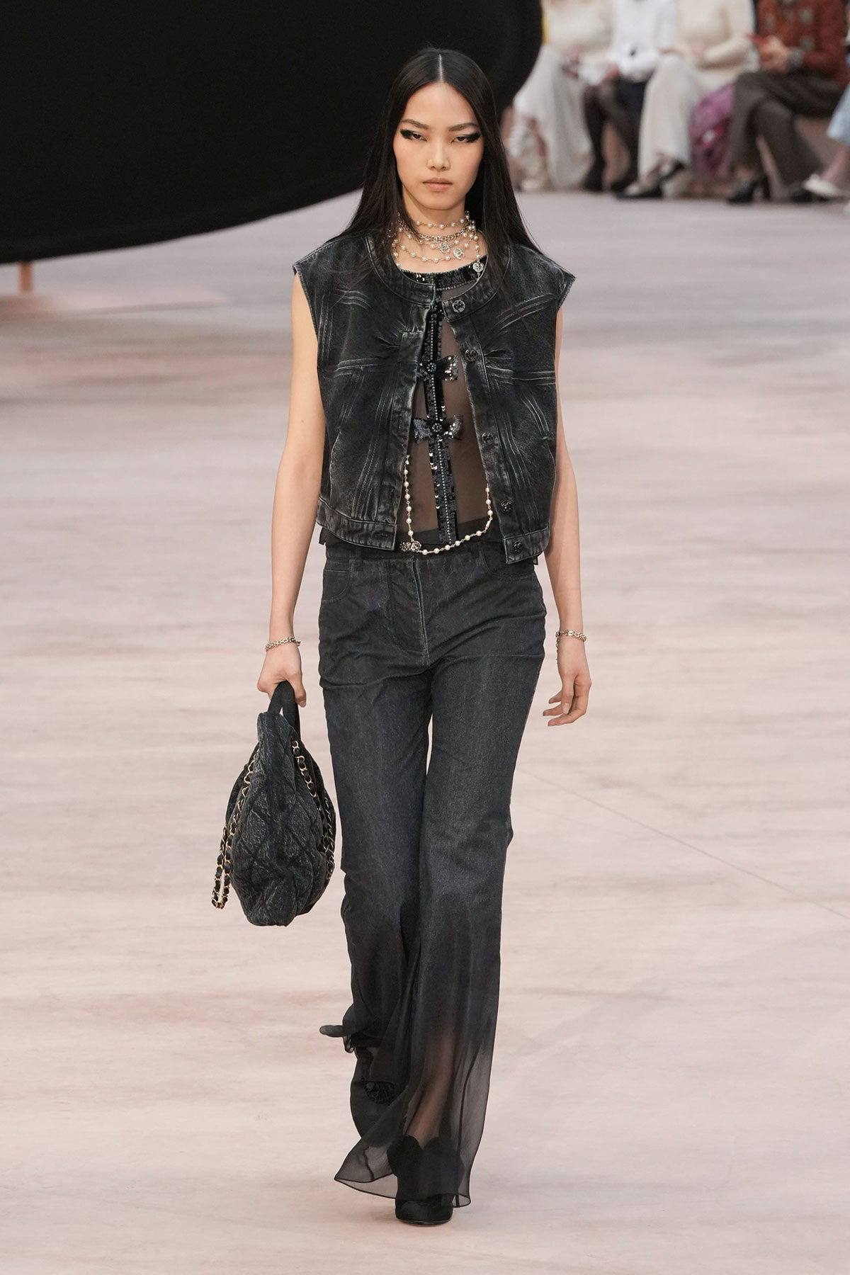 Model walking runway in black denim vest, sheer top, layered necklaces, and wide dark pants; holding a quilted black bag with chain strap.
