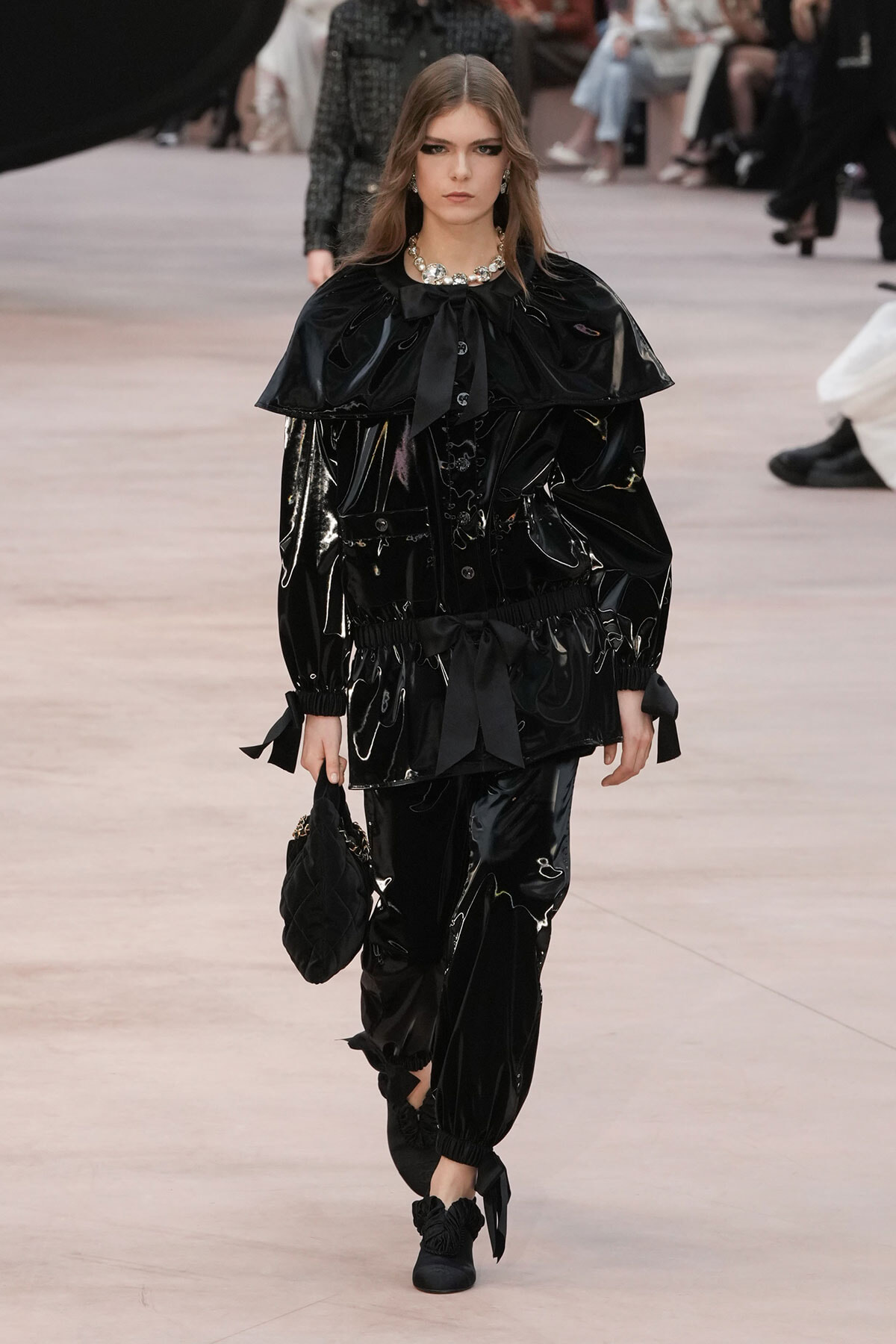 Model walking a fashion runway in a shiny black patent-look ensemble with a large cape collar and tied waist, holding a black quilted bag.