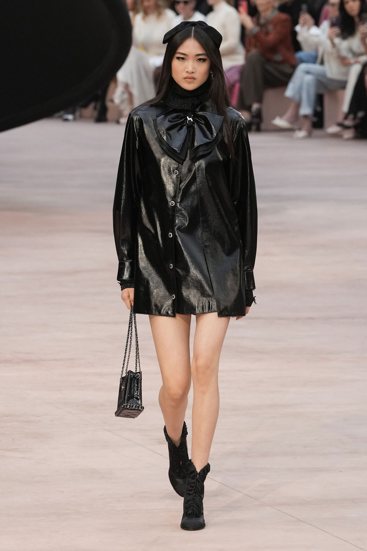Model walking the runway in a shiny black coat with a large bow at the neck and a black headband, carrying a small quilted black handbag on a chain.
