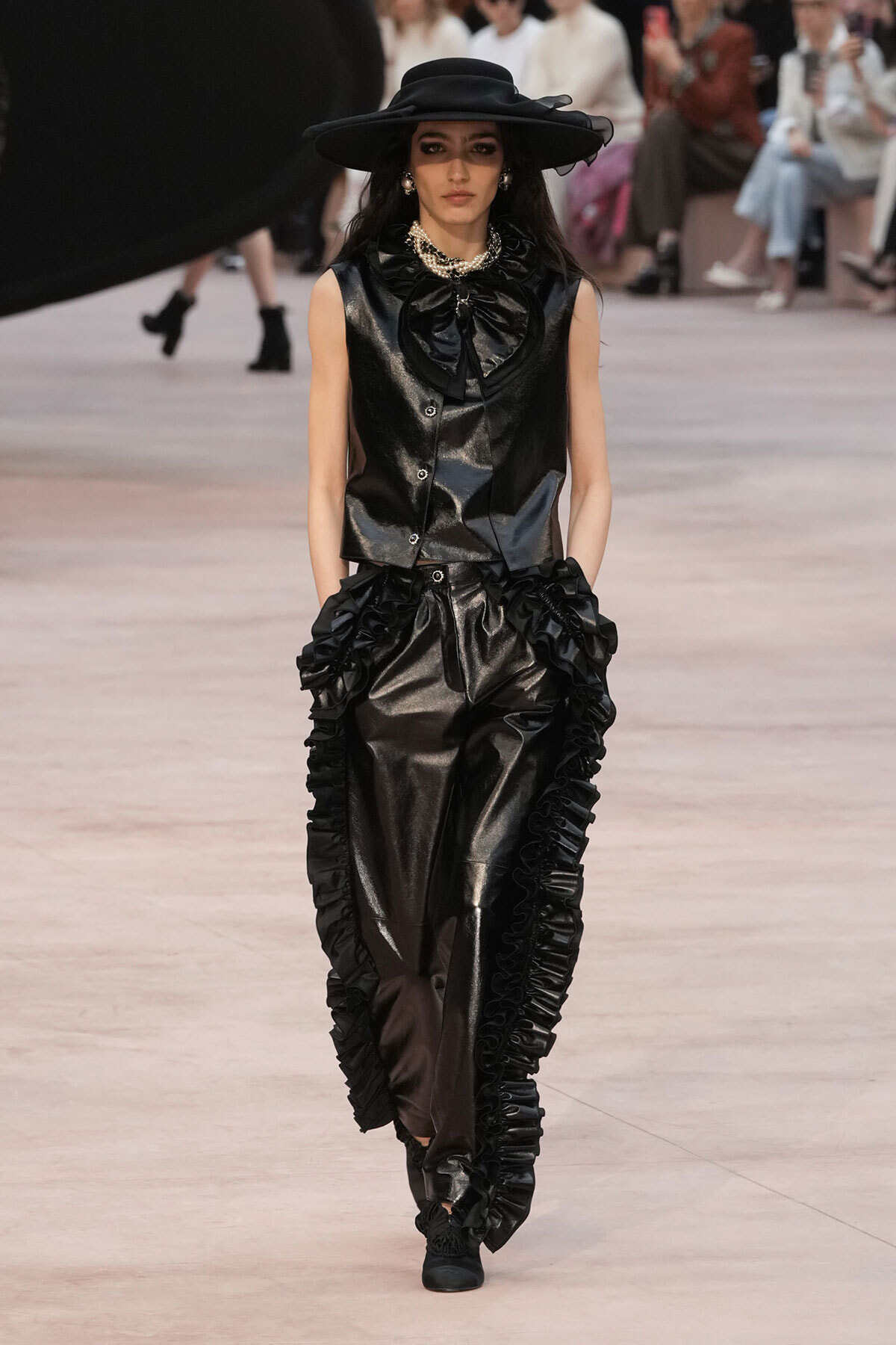 Model on a runway wearing a black glossy vest and matching wide-legged pants with large ruffles, paired with a wide-brim black hat and pearl jewelry.