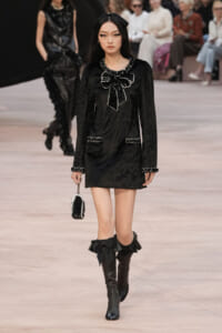 Model walks the runway in a black velvet mini dress with white beaded trim and large bow at the neckline, paired with knee-high boots tied with oversized bows and a small beaded handbag.
