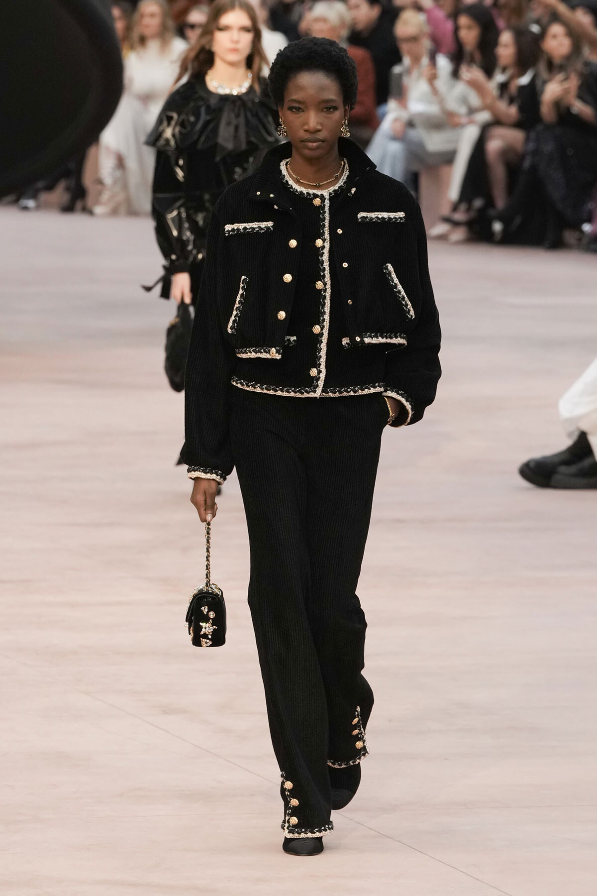 Model on a runway wearing a black textured velvet suit with gold-trimmed jacket and matching pants, carrying a small black chain-strap purse. This conveys luxury tailoring and styling for the show audience in the background.