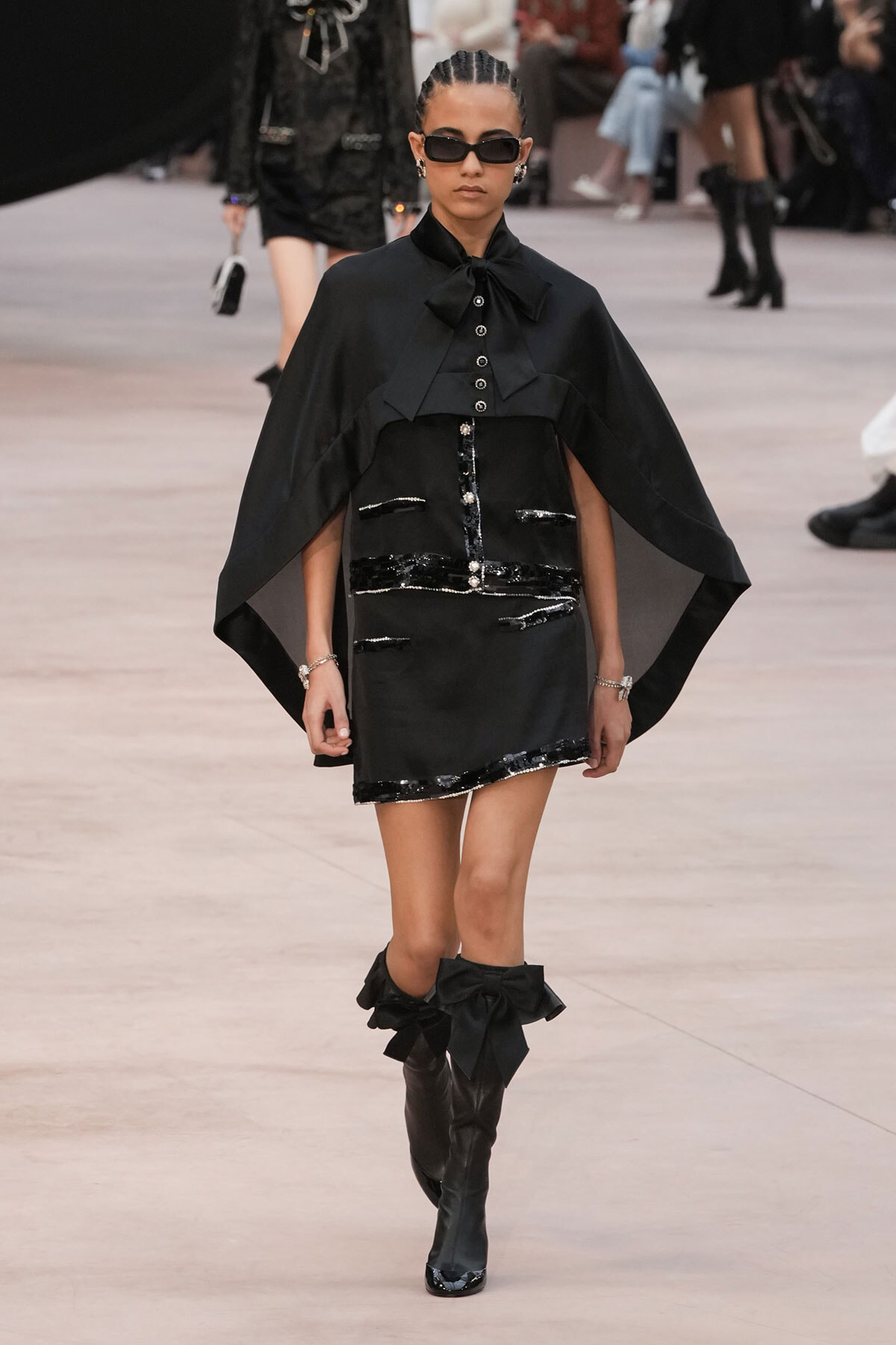 Model walking runway in a black cape-style top over a sequined black mini dress, with knee-high boots adorned by large bows and dark sunglasses.