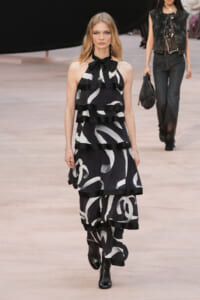 Model walking runway in black-and-white abstract print tiered dress with bow necktie and black boots.