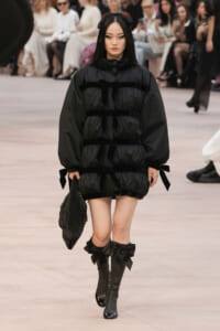 Model walking the runway in a black oversized puffer dress with ribbon ties, carrying a quilted bag with a chain strap.
