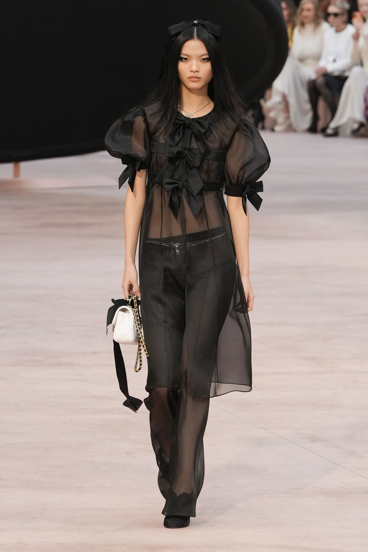 Model walking runway in sheer black dress with puff sleeves and ribbon bows, holding a small white bag with a chain strap.