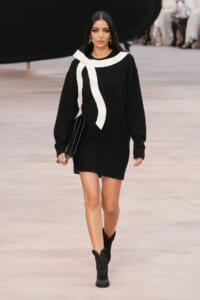 Model walking a runway in a black sweater dress with a bold white scarf detail, carrying a black clutch and wearing black boots.