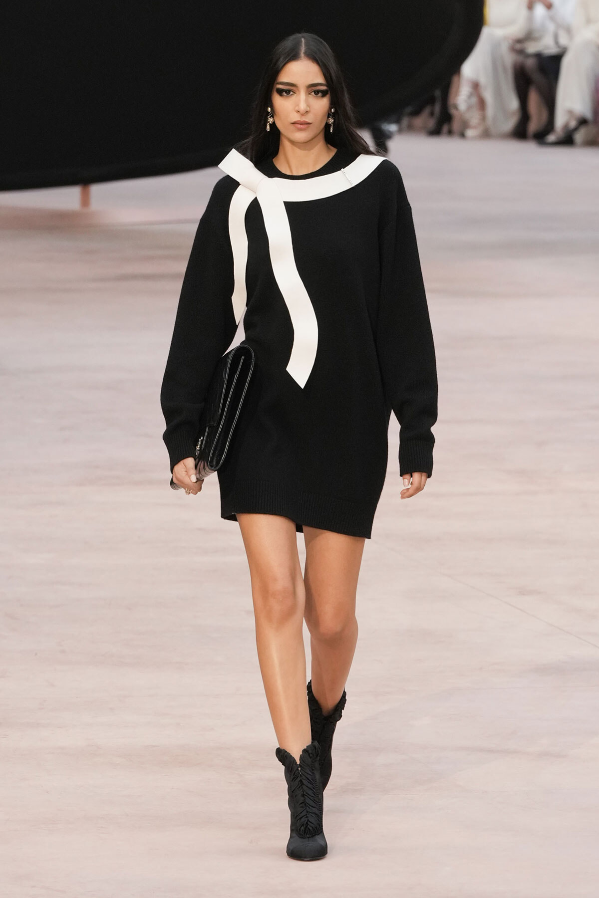 Model walking a runway in a black sweater dress with a bold white scarf detail, carrying a black clutch and wearing black boots.