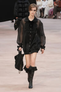 Model walks the runway in a sheer black blouse with a ruffled front, puff sleeves, and black ankle boots, carrying a black chain-strap bag.