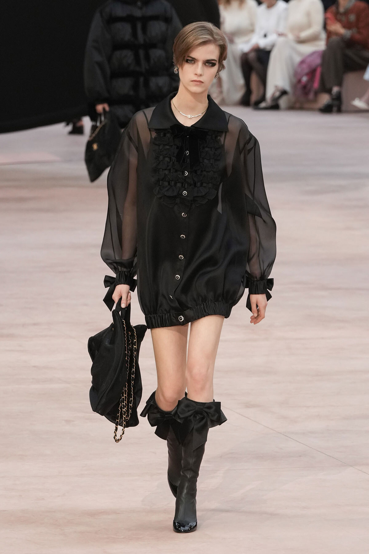 Model walks the runway in a sheer black blouse with a ruffled front, puff sleeves, and black ankle boots, carrying a black chain-strap bag.